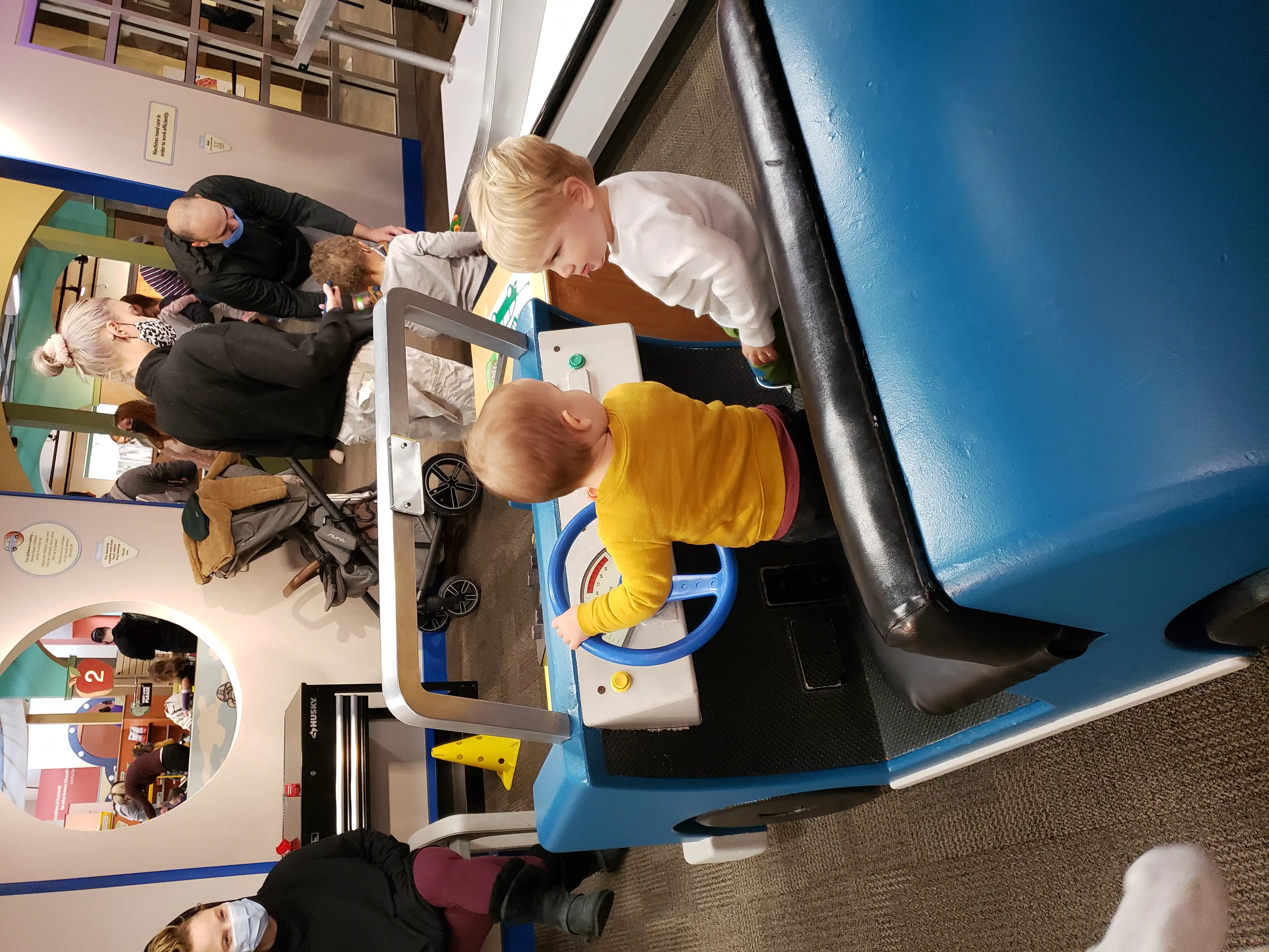 Royal in a play car next to another boy in a crowded room. They're staring at one another.