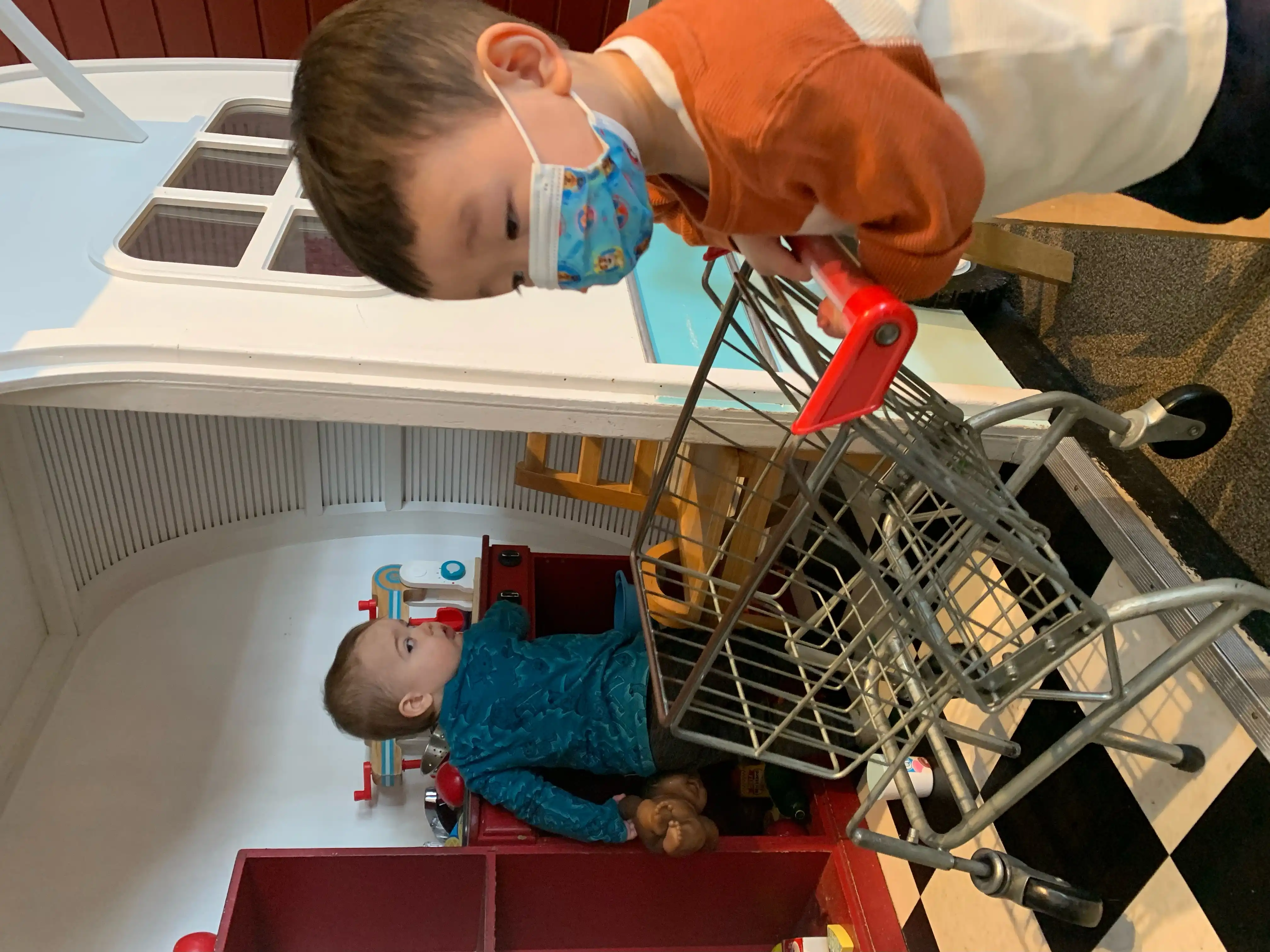 Royal in a toy house looking back at Nikki pushing a miniature shopping cart inside.