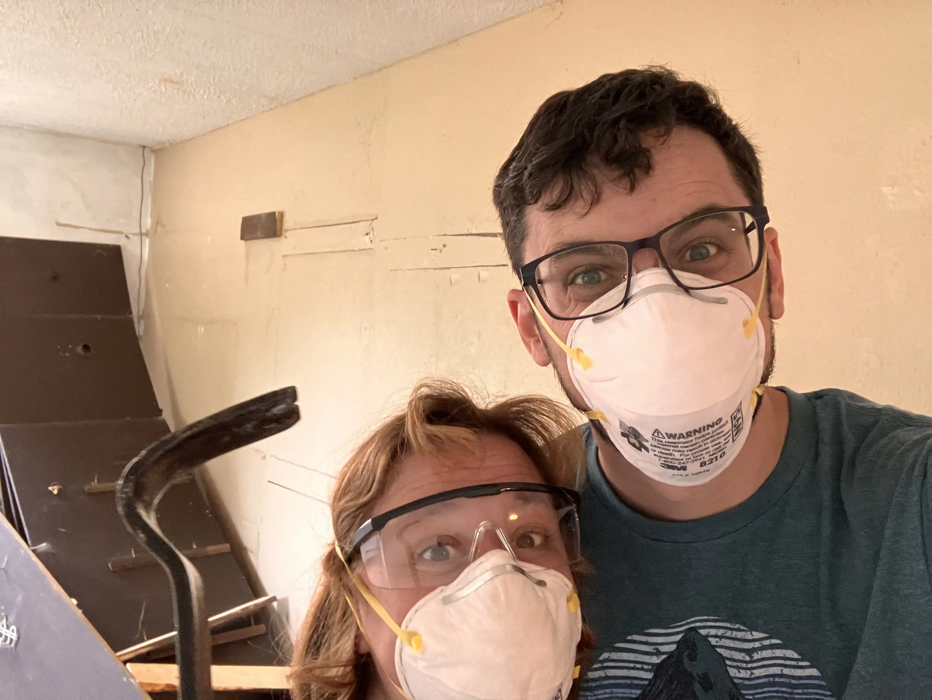 Alex and Amie with painter's masks holding a crowbar in the basement, where all the shelves are tossed around the room.