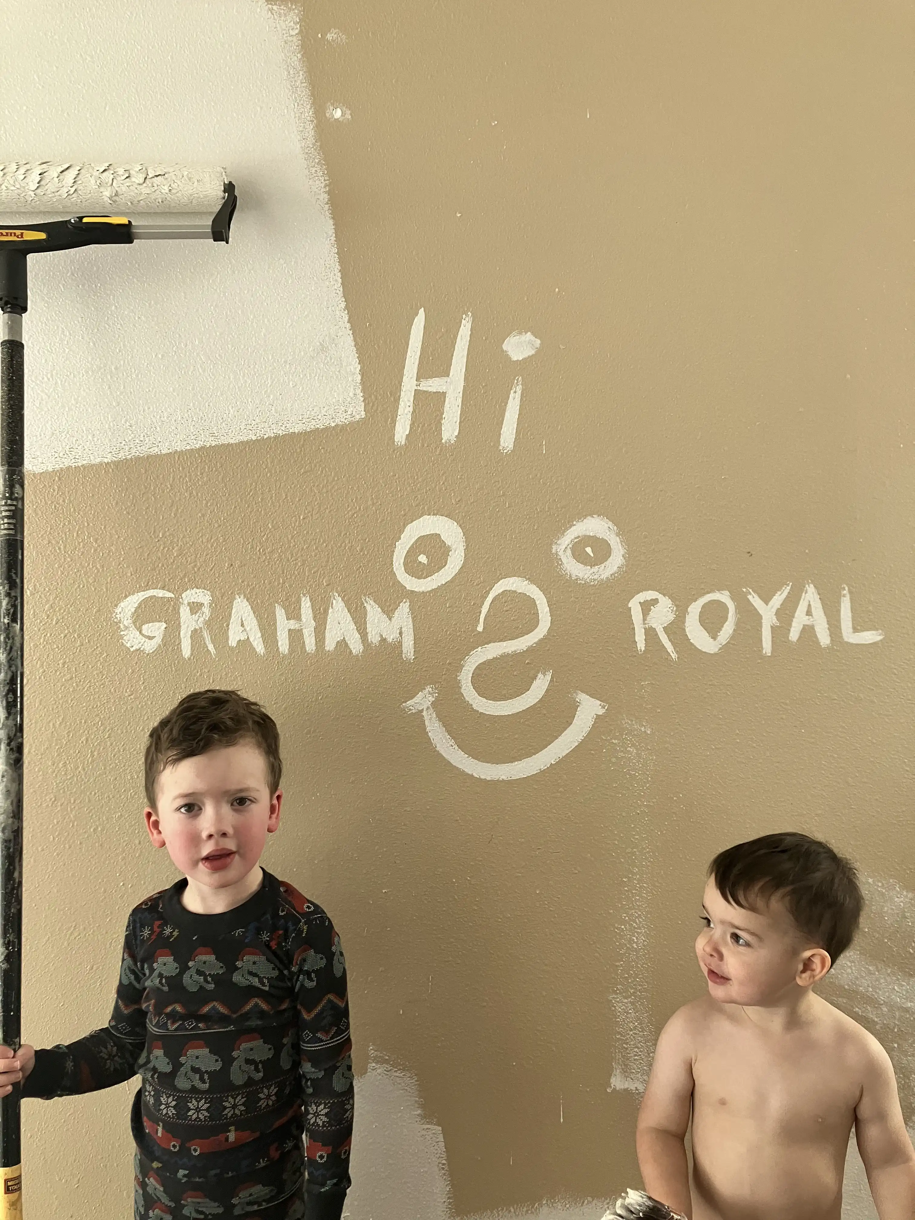 Graham holding a paint roller next to Royal. On the wall behind are their names and a smiley face in white primer over the brown wall color.