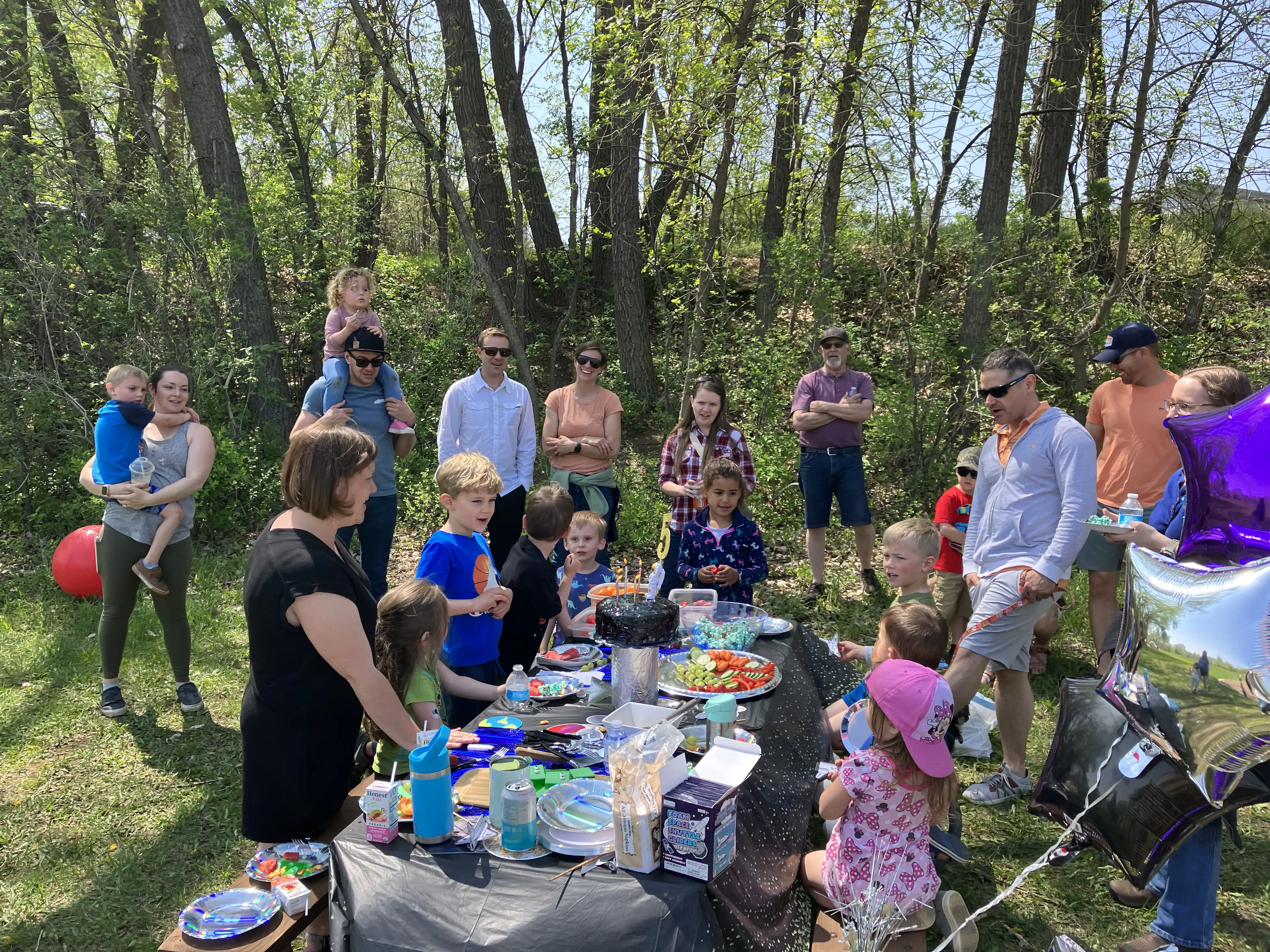 All the party goers around the birthday table. In order from left-to-right: Gina and Reese, Amie, Nate with his daughter on his shoulders, Flora and Augustus at the table, William and his wife behind, with Graham and Kyle at the table, Cora with Flora's mom behind, Grandpa Tom, then Lily, Royal, and Jorgen seated at the table, Levi in sunglasses behind Uncle Tom, then John and finally Teresa. Whew!