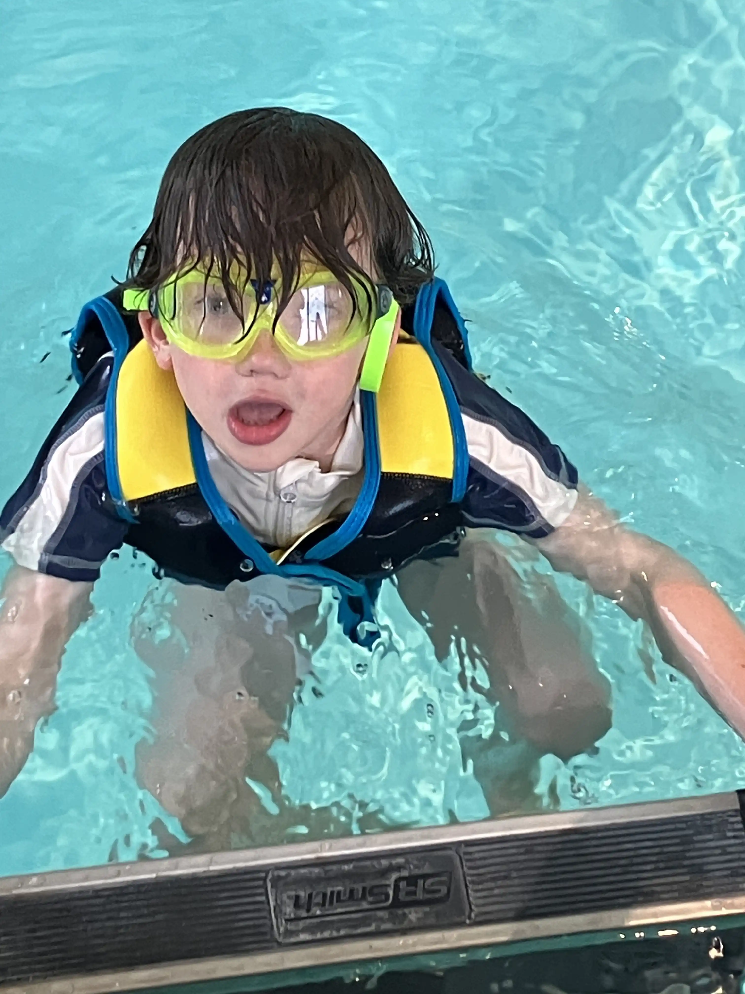 Graham wearing swim goggles and a life vest at the pool's edge